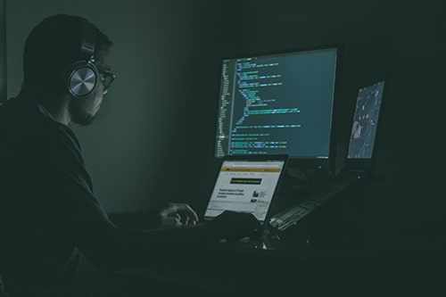 Programador trabalha em seu notebook e monitor, em um quarto escuro