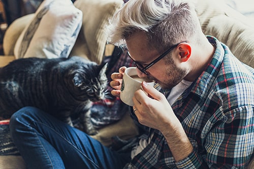 Homem tomando café no sofá ao lado de seu gato.